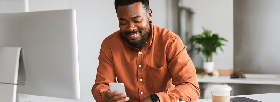 Man wearing an orange shirt using a cell phone next to a desktop computer Man wearing an orange shirt using a cell phone next to a desktop computer