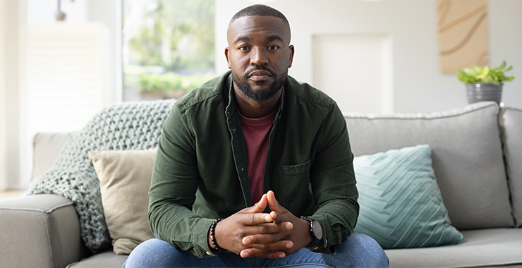 Man wearing a green jacket sitting on a gray couch looking at the camera