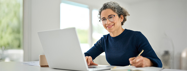 Woman wearing a blue shirt writing while smiling at her laptop