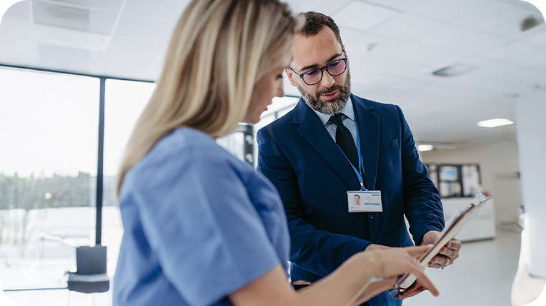 Man wearing a blue suit holding a clipboard while talking to a woman wearing blue scrubs